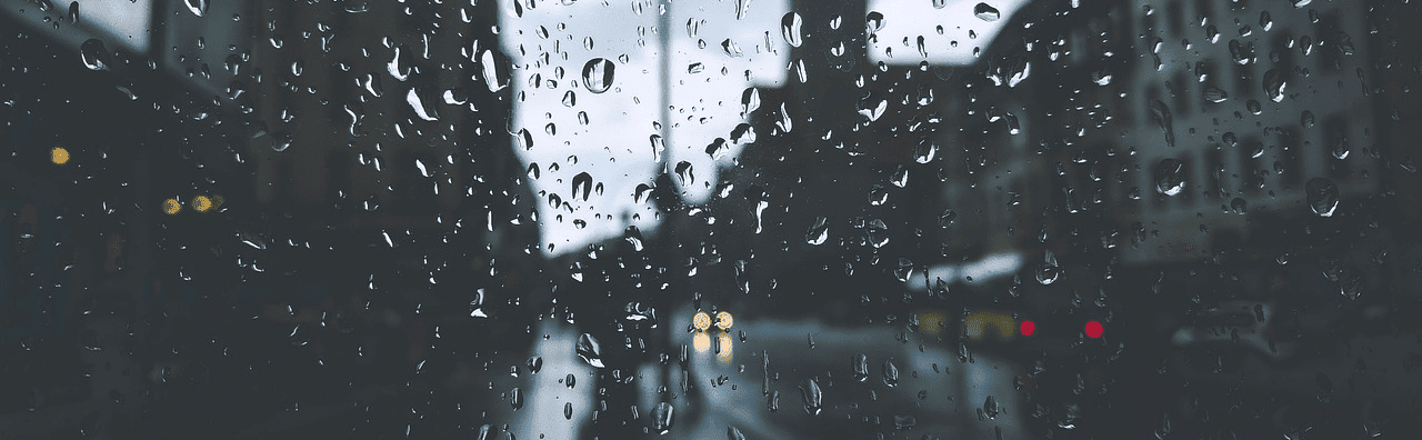 Rain on a camera lense pointing towards the streets. Shadows and clouds making it dark, 1 car and buss in the distance.